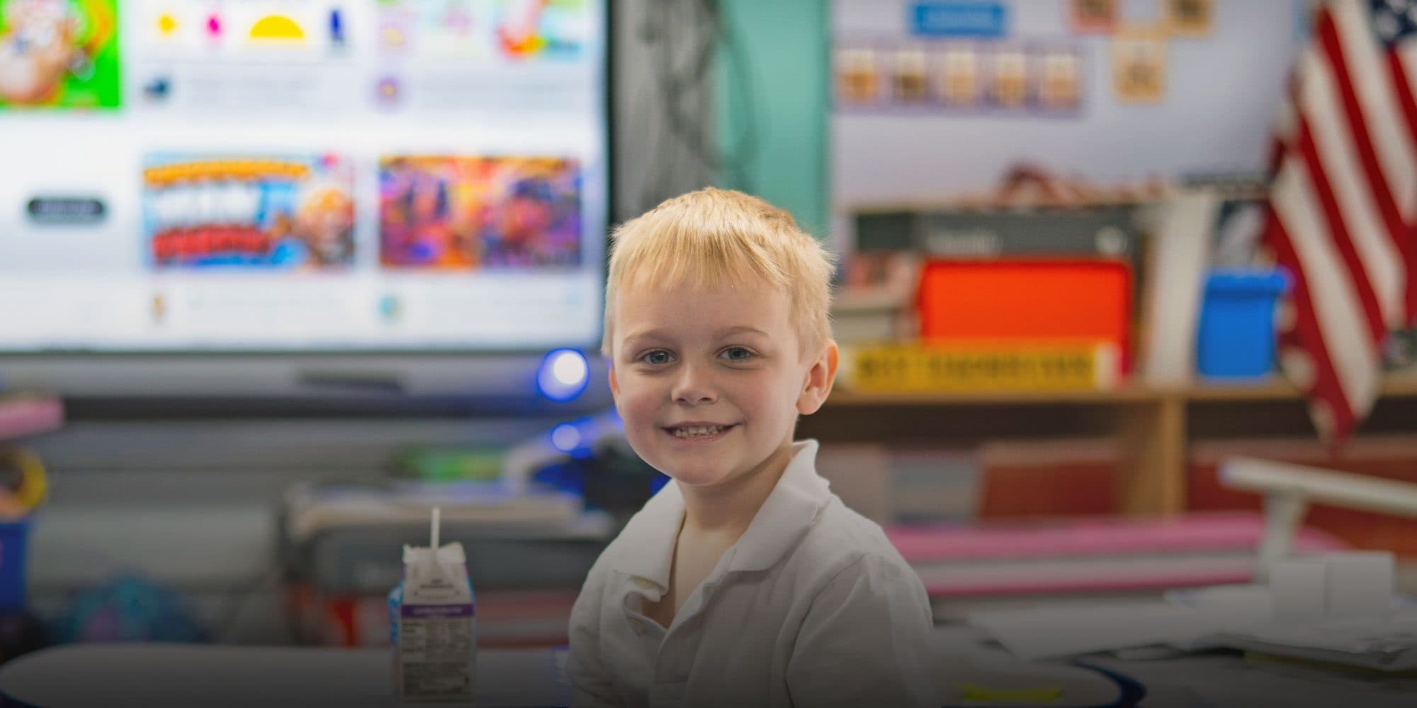 Smiling student at a classroom desk in front of a large display screen.
