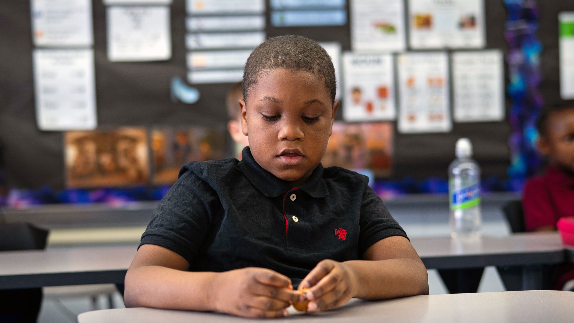 Student peeling a small orange at a classroom desk.